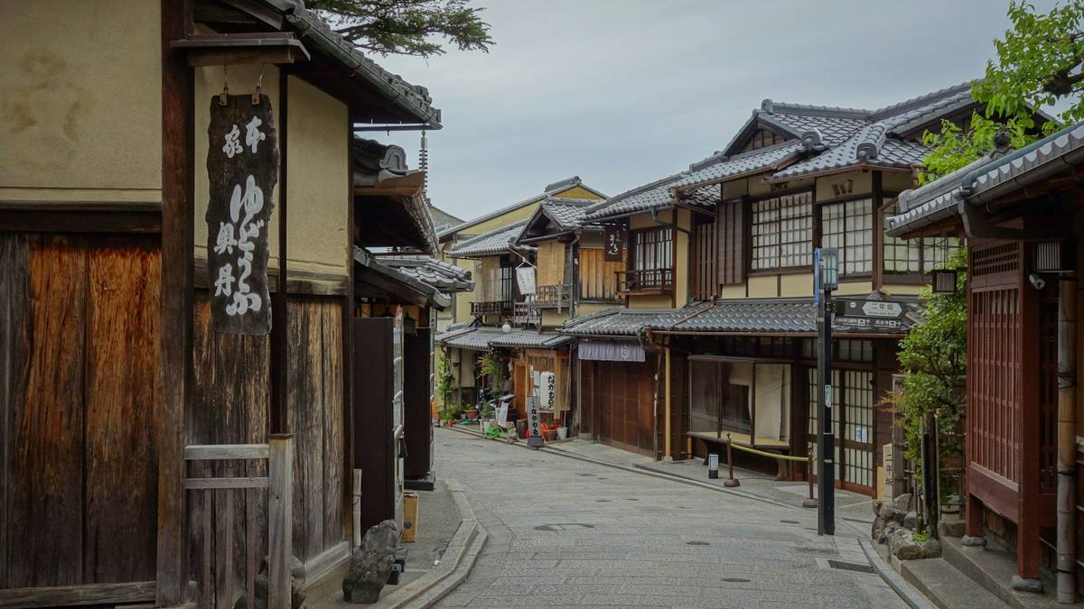 Smal gate i Kyoto mellom toetasjes trehus med mørke bordkledninger, gittervinduer og et butikkforheng med japanske skrifttegn