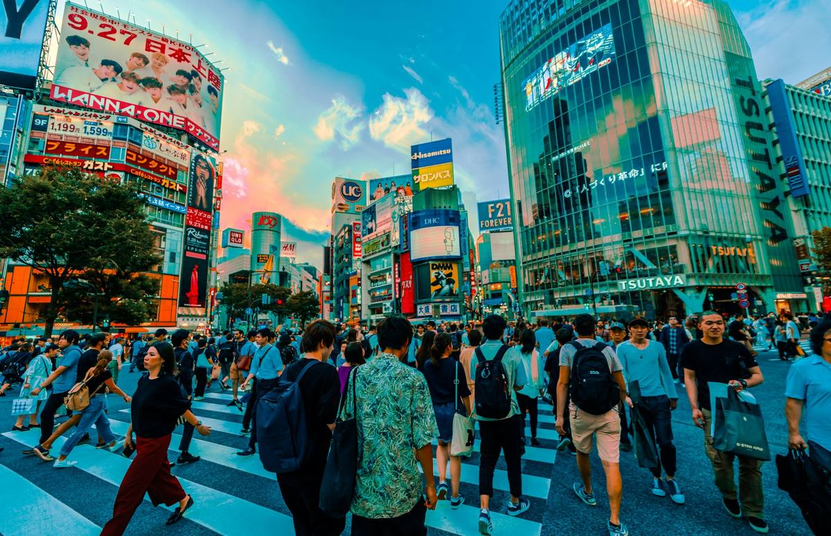 Folkemengde som krysser fotgjengerfeltet i Shibuya, omgitt av høye fasader dekket av lysende reklameskjermer og butikkskilt