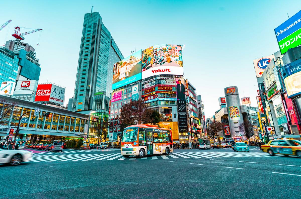 Fotgjengerkrysset i Shibuya i skumringen med sebrastriper, en liten rød buss midt i krysset og reklameskjermer på husveggene rundt