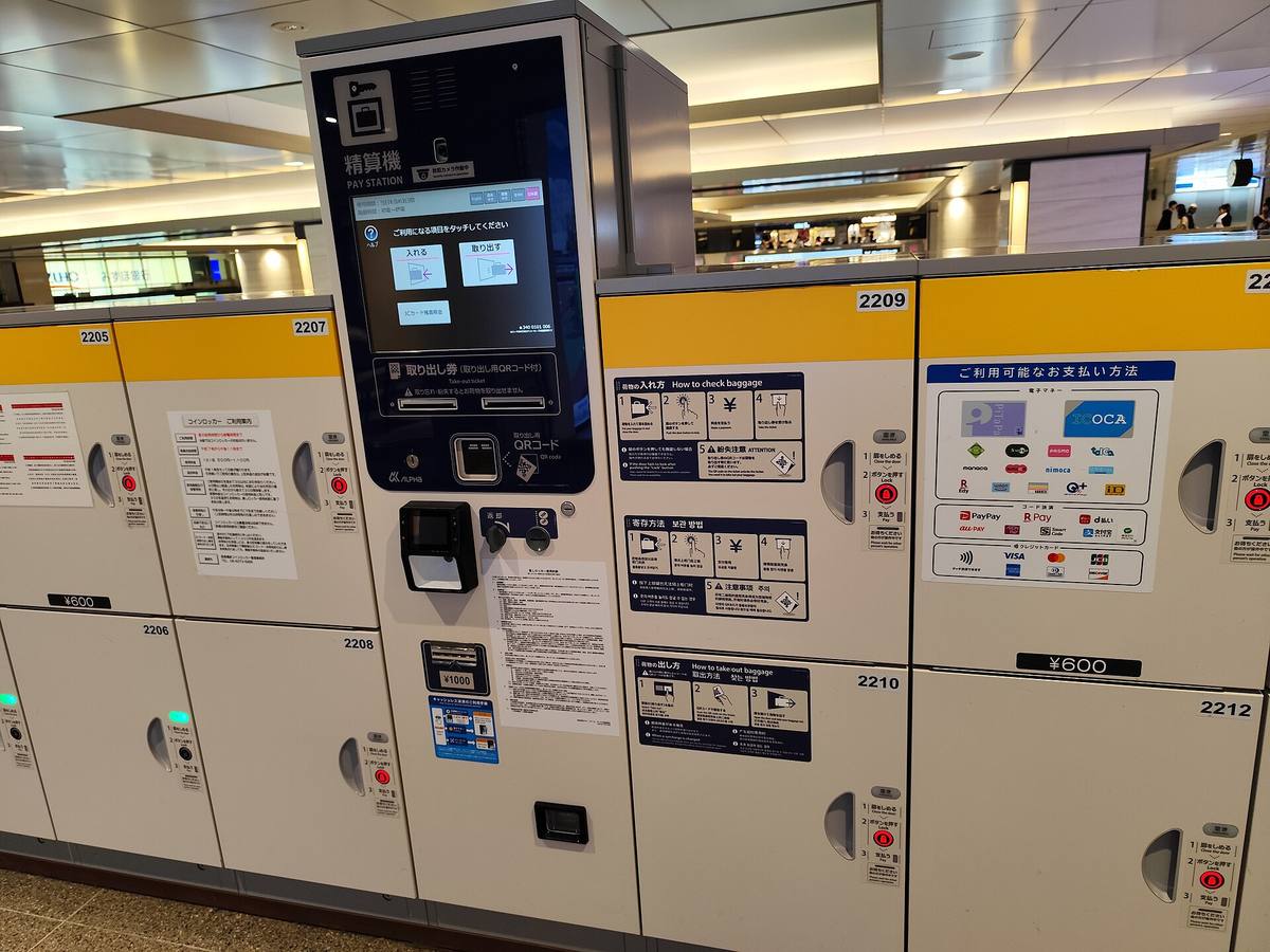 Bank av hvite bagasjebokser med gul topp på en jernbanestasjon, med nummererte dører, betalingsautomat i midten og bruksanvisning på japansk og engelsk