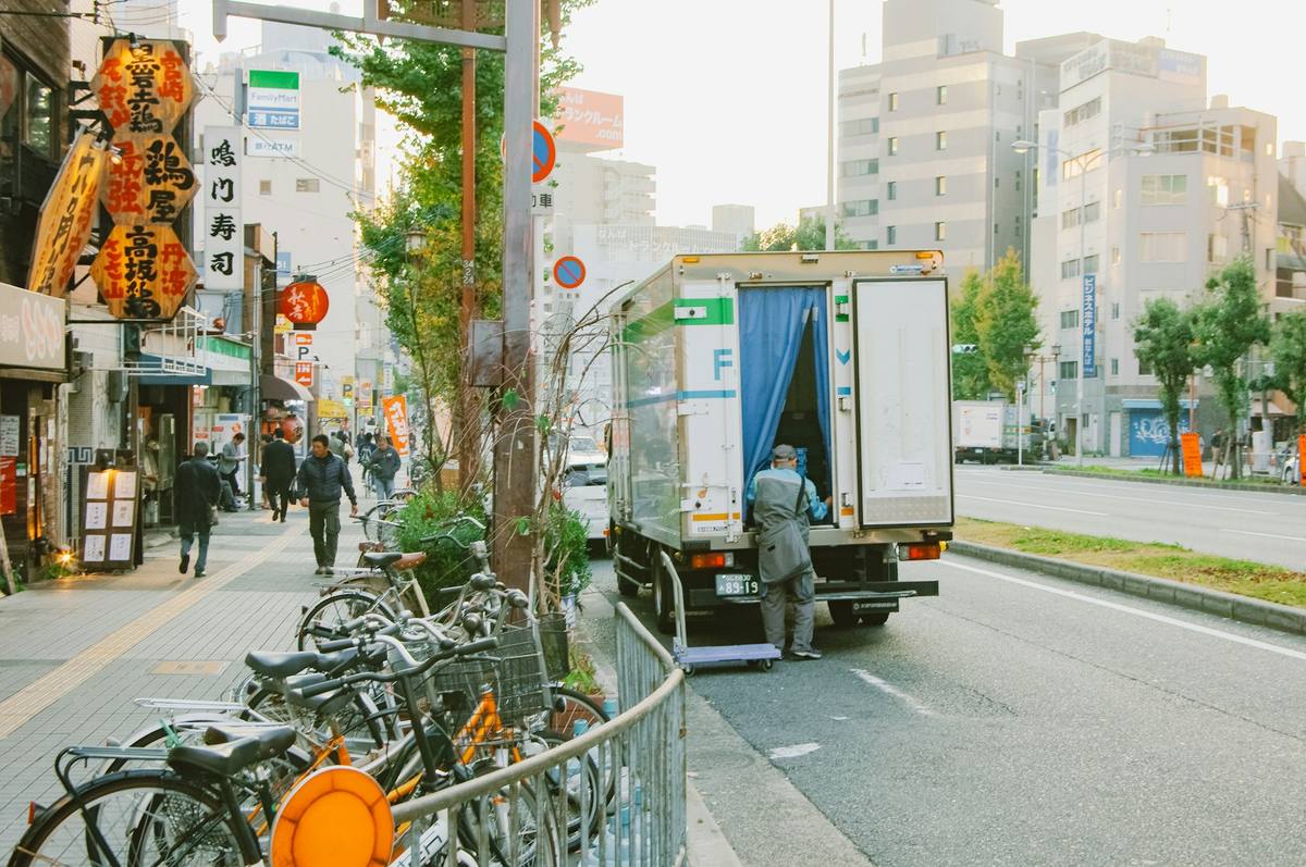Hvit varebil med åpne bakdører og blått forheng står i en japansk bygate mens en budbilsjåfør med trillebord laster ut, med sykler og butikkskilt langs fortauet