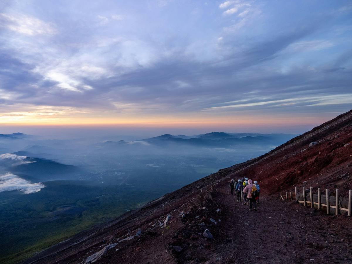 Rekke av fjellvandrere med hjelm og ryggsekk på den røde grusstien høyt på Fuji ved daggry, med skylag og blå fjellrygger langt nede