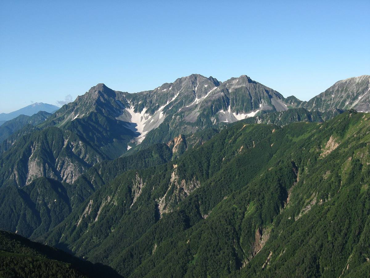 Hotaka-massivet i Japanalpene med snøflekker i klippene og tett grønn skog nedover fjellsidene, under klar blå himmel