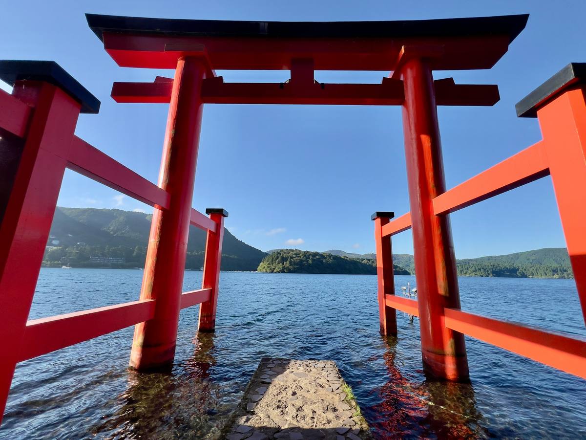 Rød torii-port som står ute i vannet ved Ashi-sjøen i Hakone, sett tett på nedenfra, med skogkledde åser på motsatt side