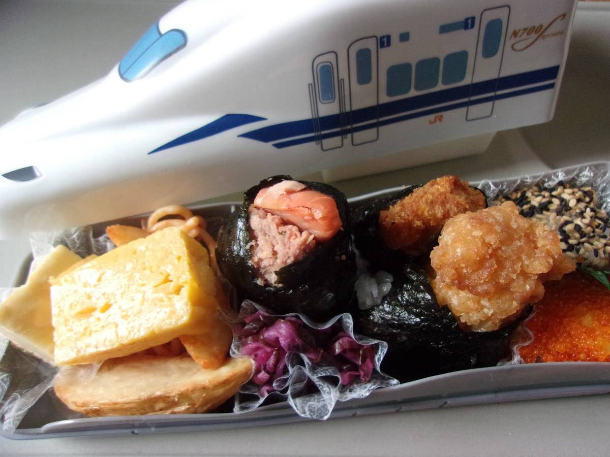 Åpen togmatboks med omeletter, risballer i tang, frityrstekt kylling og syltede grønnsaker, med lokket formet som et hvitt shinkansen-tog bak