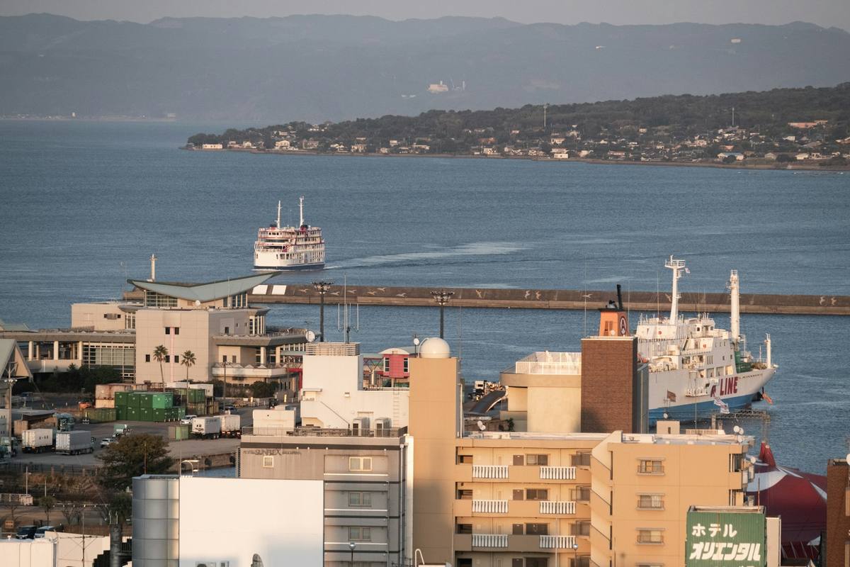 Hvit passasjerferge på vei inn mot havnen bak moloen, med en større bilferge til kai og bykvartaler langs sjøkanten