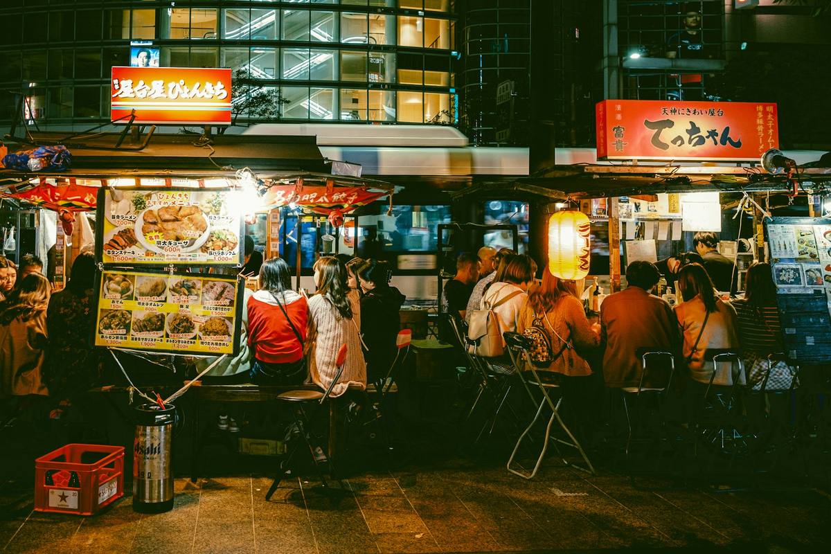 Opplyste yatai-matboder på fortauet en kveld, med gjester på klappstoler langs disken og menyplakater på japansk