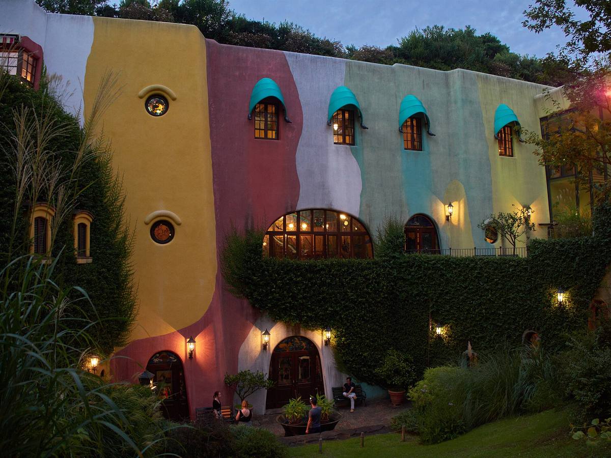 Fasaden på Ghibli-museet i skumringen, med bølgete felt i gult, rosa og grønt, turkise buer over vinduene og eføy og gress rundt inngangene