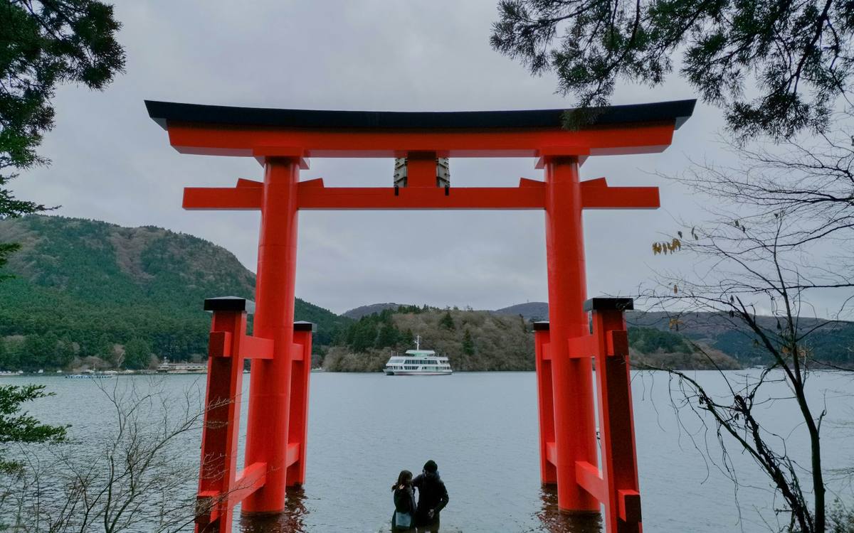 Stor rød torii-port som står ute i en fjellsjø, med skogkledde åser og en turbåt i bakgrunnen