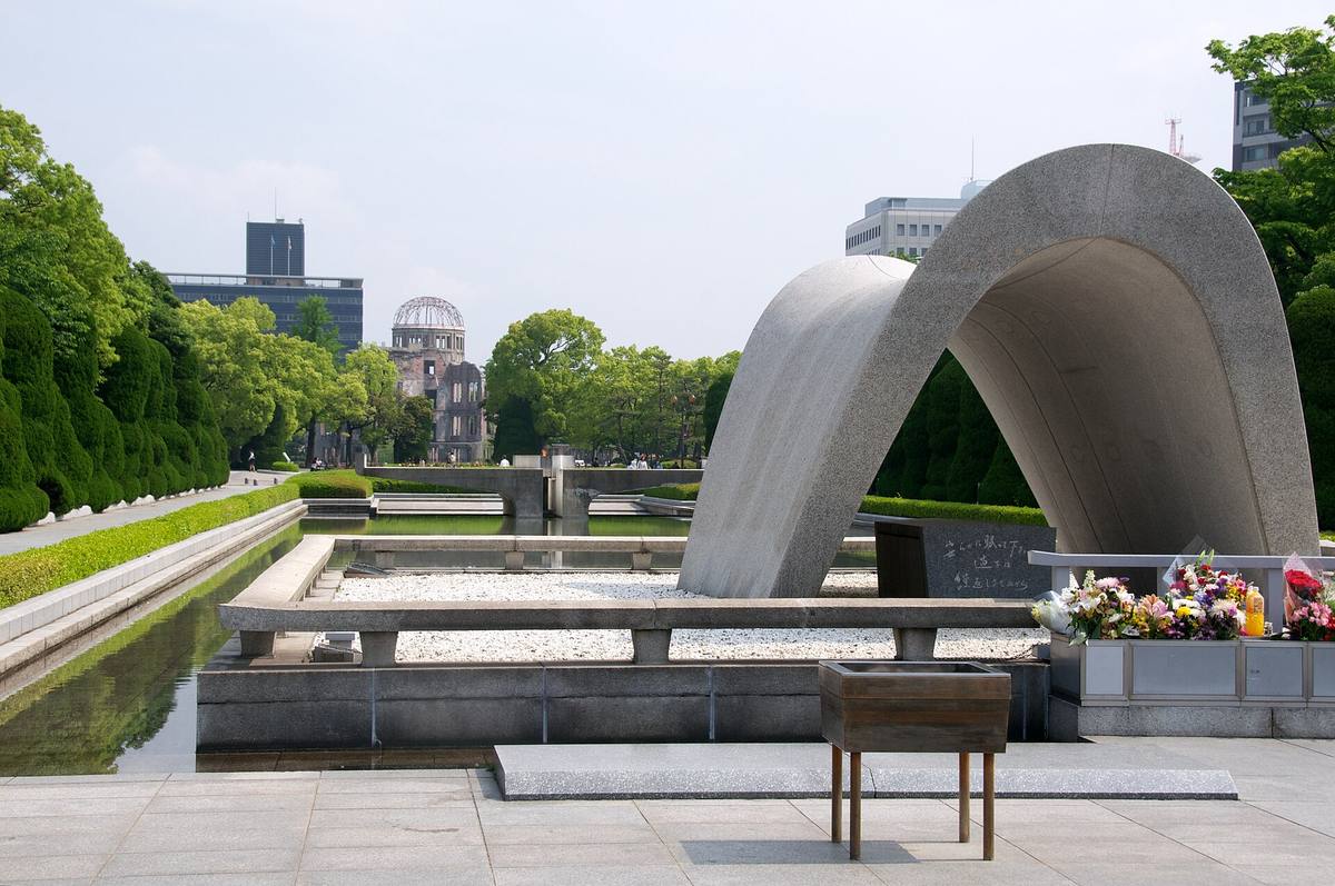 Kenotafen i Fredsparken i Hiroshima, en buet steinhvelving med A-bombekuppelen synlig gjennom åpningen og blomster ved foten