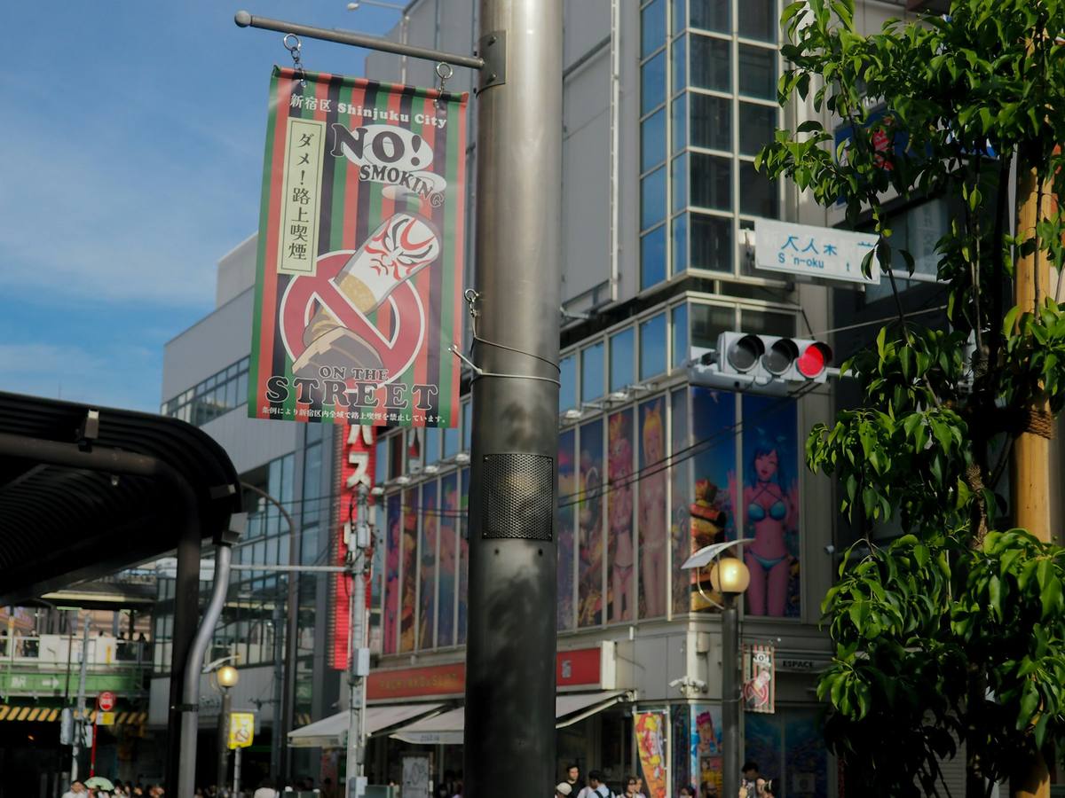 Banner fra en bydel i Tokyo hengt på en lyktestolpe med en overstrøket sigarett og teksten om at røyking på gaten er forbudt, med glassfasader og et lyskryss bak