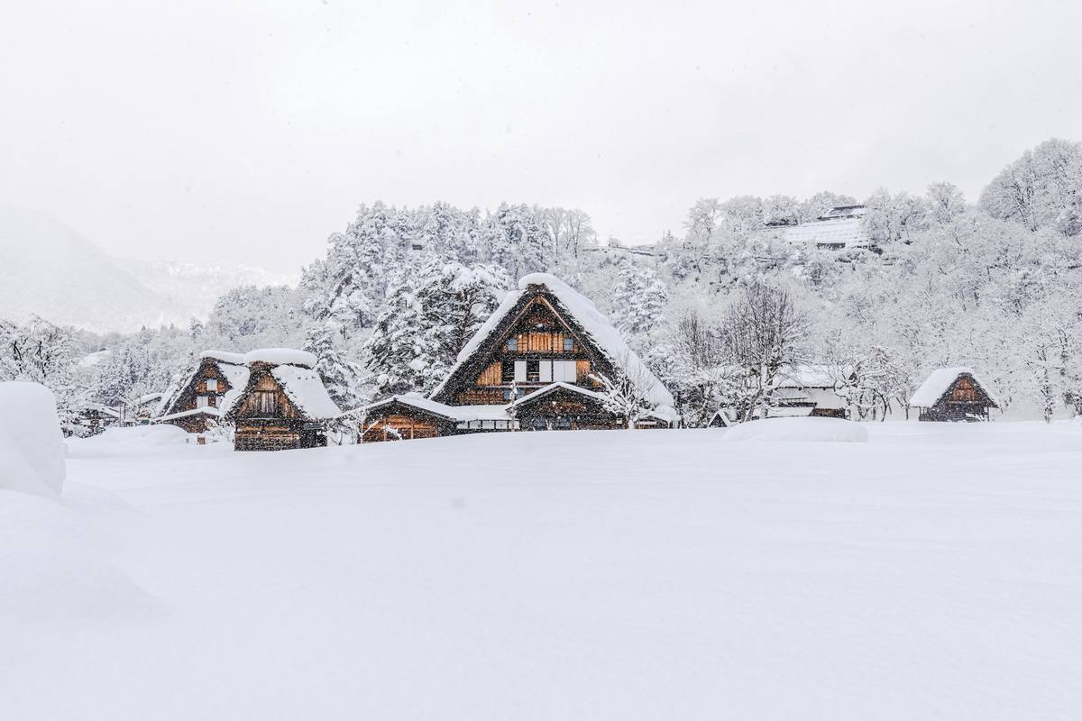 Gårdshus med bratte halmtak nesten begravd i snø, med snødekt skog bak og fallende snøfnugg i luften
