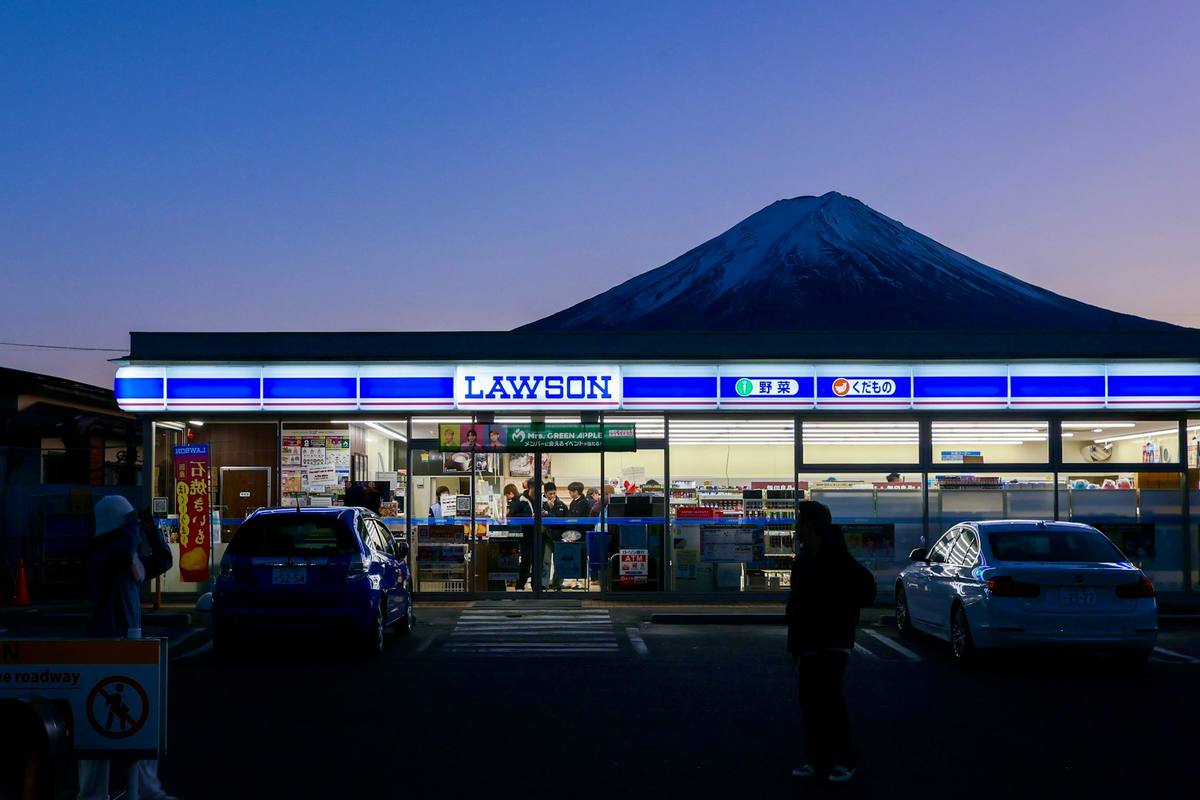 Opplyst Lawson-nærbutikk i skumringen med Fuji som reiser seg rett bak taket, og et par biler på plassen foran