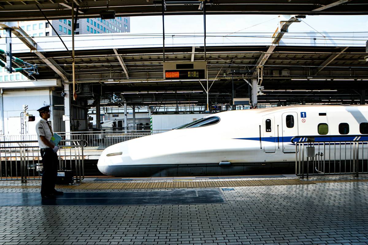 Shinkansen med lang, spiss nese står inntil perrongen mens en konduktør i uniform venter ved sporet under et oransje avgangsskilt på japansk