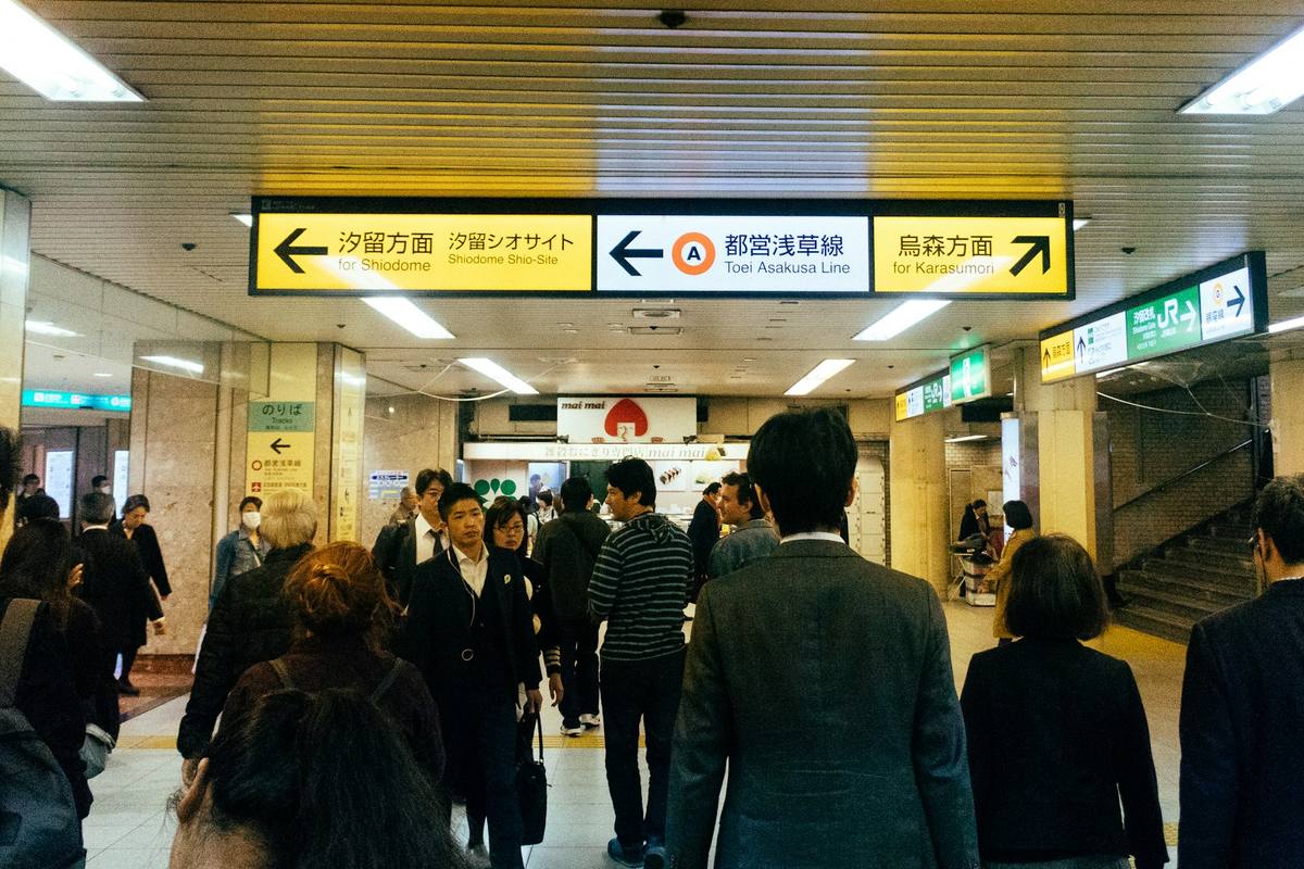 Underjordisk stasjonshall med gule retningsskilt på japansk og engelsk, der pendlere i mørke dresser går mot utgangene