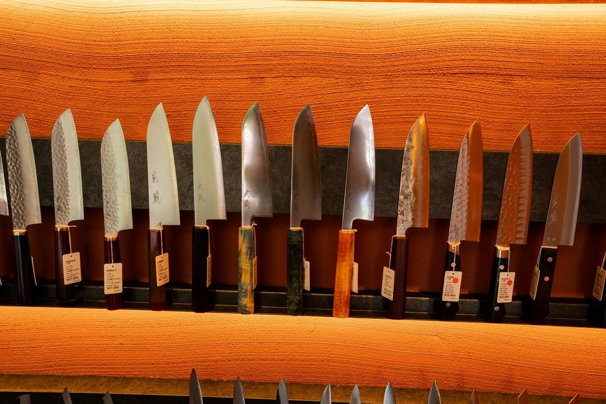 Rekke japanske kjøkkenkniver med hamrede blad og håndtak i ulike tresorter, hengt på en trevegg med prislapper