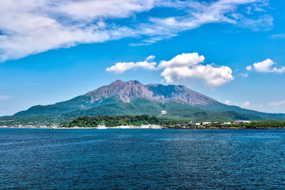 Vulkanen Sakurajima sett over blått sjøvann fra fergen, med grønnkledd fot, grå vulkantopp og hvite skyer mot blå himmel