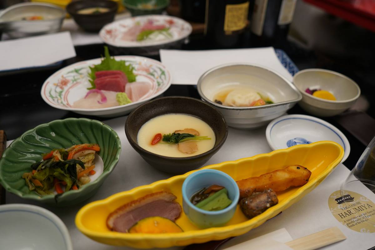 Måltid med mange små retter i ulike skåler og fat på et bord: sashimi, dampet eggestand, syltede grønnsaker og en gul avlang skål med fisk