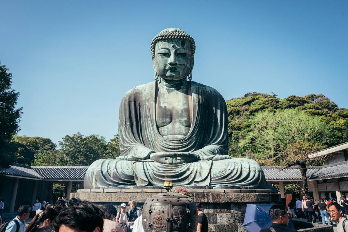 Den store bronsebuddhaen i Kamakura sittende i lotusstilling på en steinsokkel, med skogkledd ås bak og besøkende på plassen foran