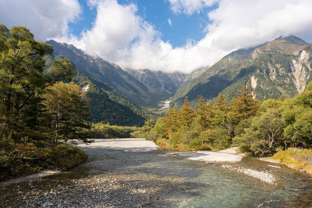Den grunne Azusa-elven renner over lys grus i en bred fjelldal med skogkledde lier og skyer som legger seg over de grå toppene