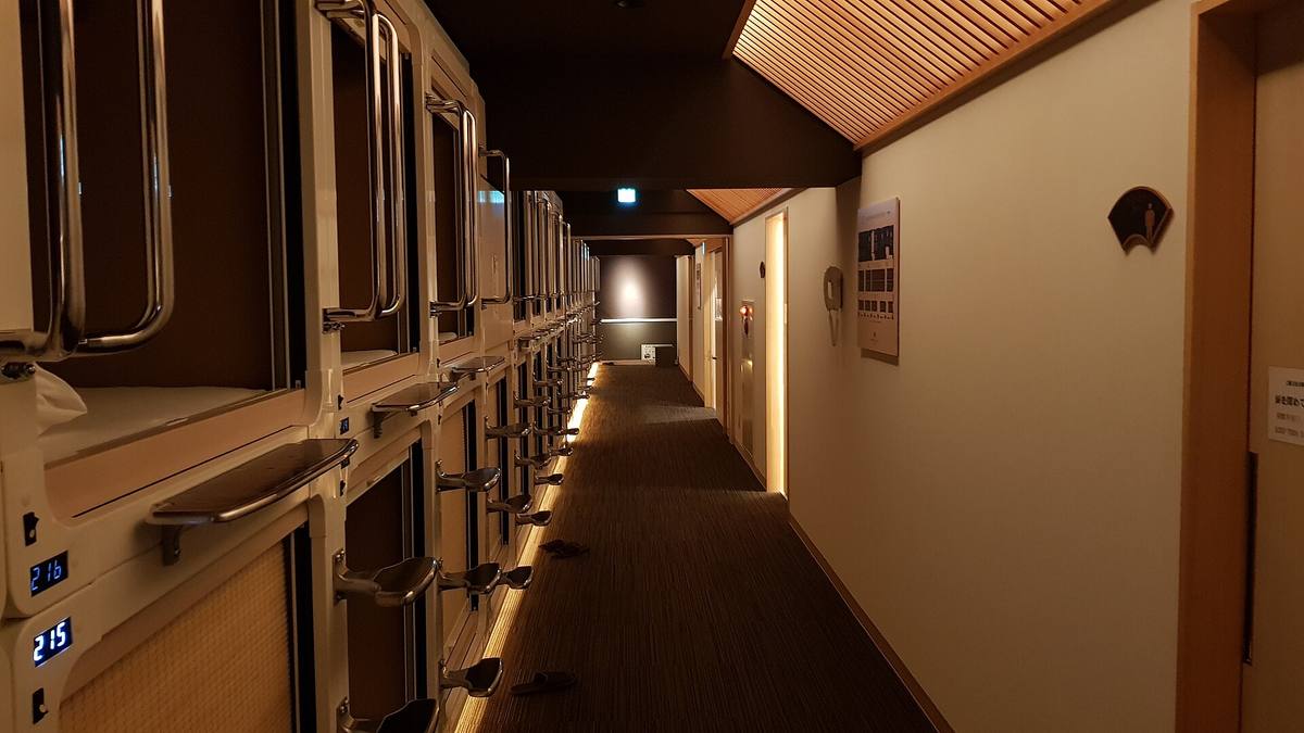 Korridor på kapselhotell med to etasjer av kapsler bak åpne luker, nummererte plasser og dempet lys langs veggen