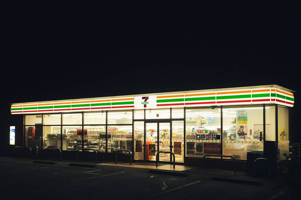 Opplyst 7-Eleven-døgnbutikk med fulle hyller bak glassfasaden og tom parkeringsplass foran i nattemørket