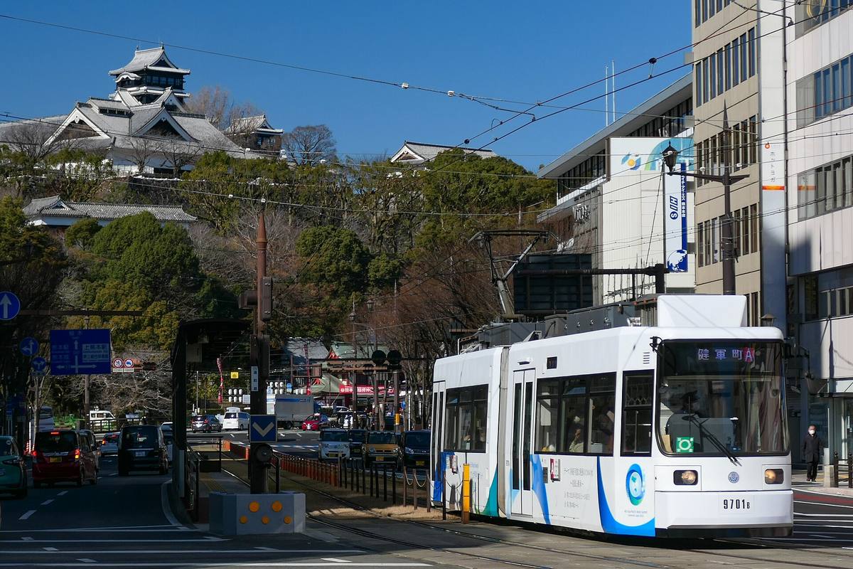 Hvit og blå trikk i gaten i Kumamoto, med borgtårnet på Kumamoto-slottet hevet over trærne i bakgrunnen
