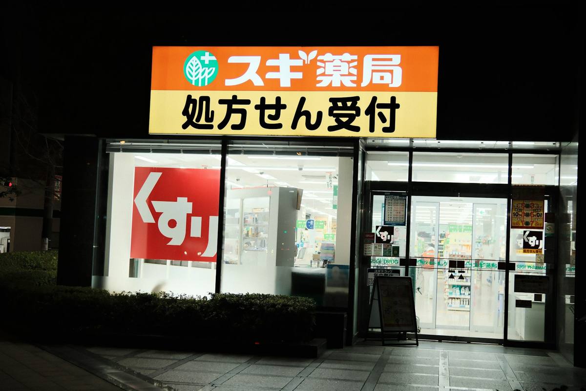 Fasaden på et japansk apotek om kvelden med opplyst oransje og gult skilt over inngangen, store japanske skrifttegn for resept og medisin i vinduet og fylte varehyller innenfor glassdørene