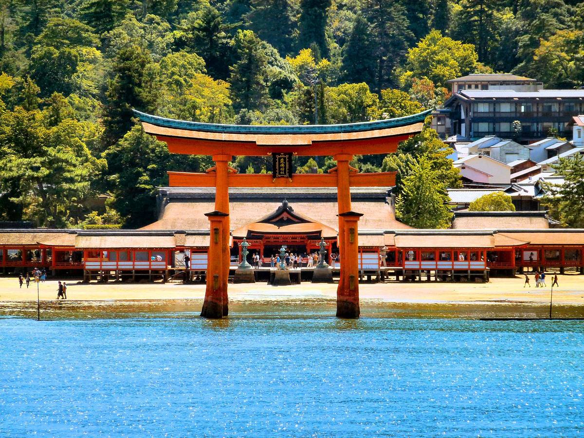 Den store vermiljonrøde torii-porten ved Itsukushima som står ute i vannet, med helligdommens bygninger og skogkledd fjell bak