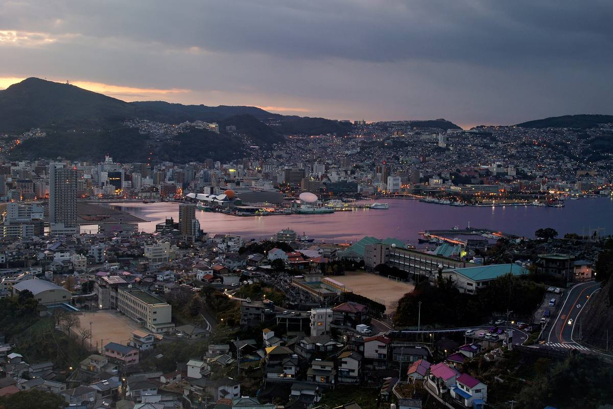 Nagasaki i skumringen sett ovenfra, med tett bebyggelse i åssidene rundt den rosa-speilende havnebukta og mørke fjell i horisonten
