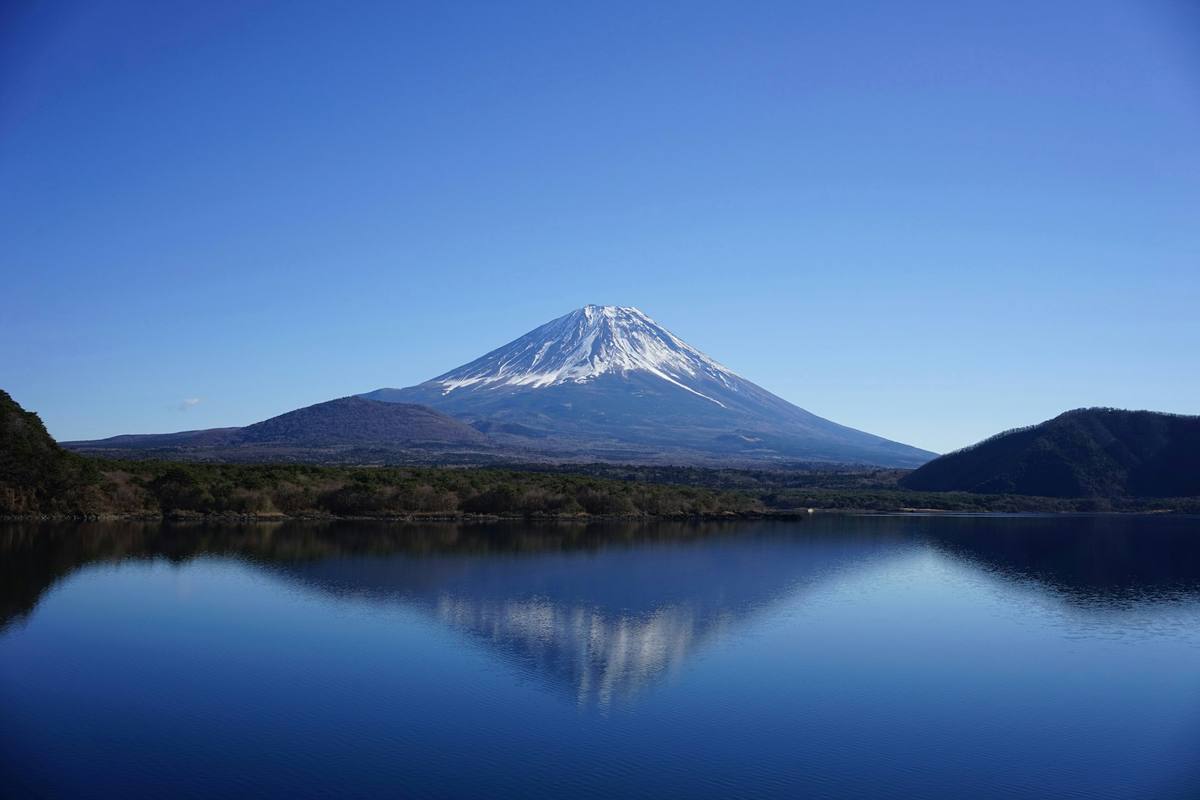 Den snødekte Fuji-kjeglen speiler seg i et blikkstille innsjøvann under klar blå himmel, med skogkledde odder på hver side