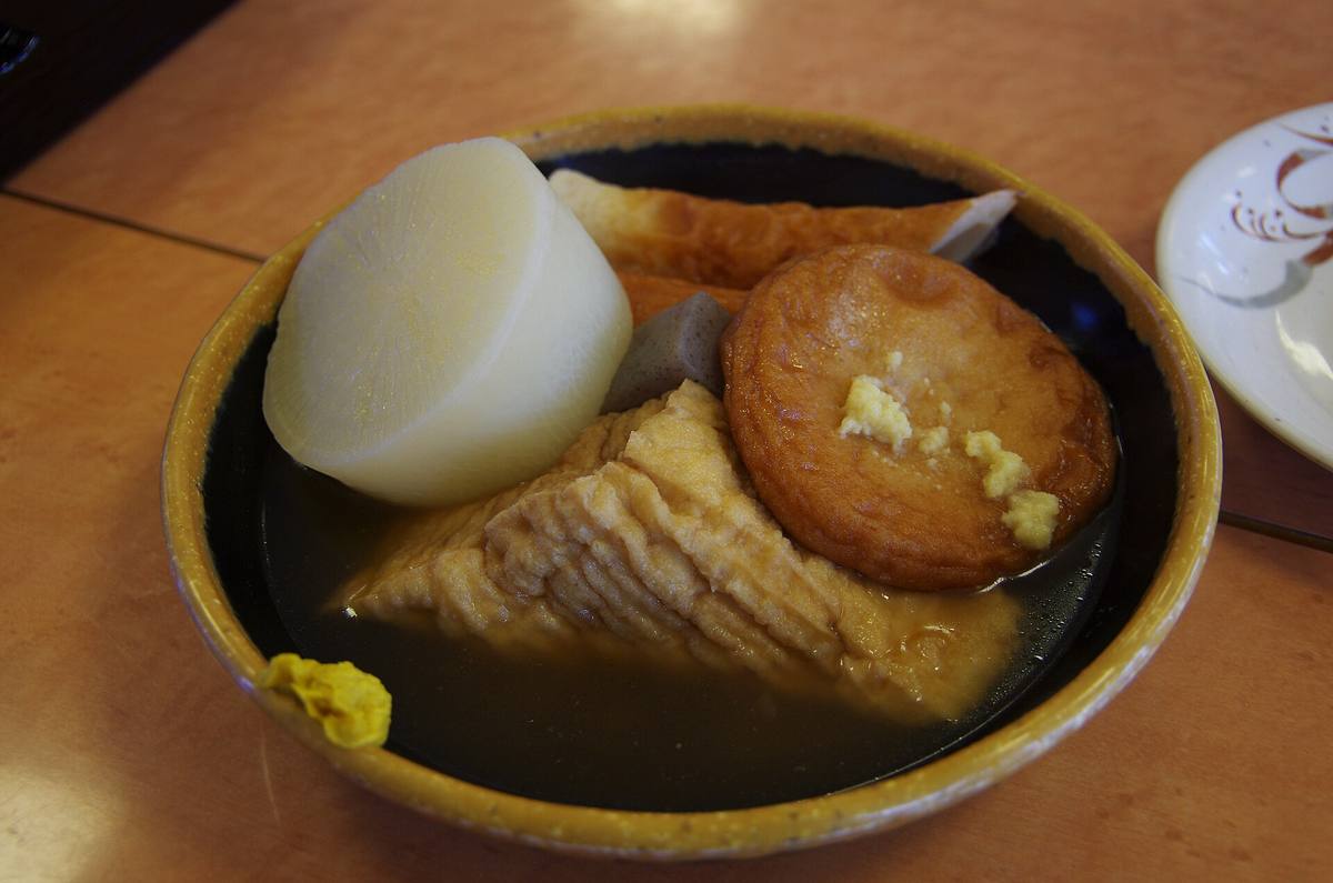 Skål med lys kraft der en stor bit kokt daikon, en trekant frityrstekt tofu og en rund fiskekake ligger, med en klatt gul sennep på kanten