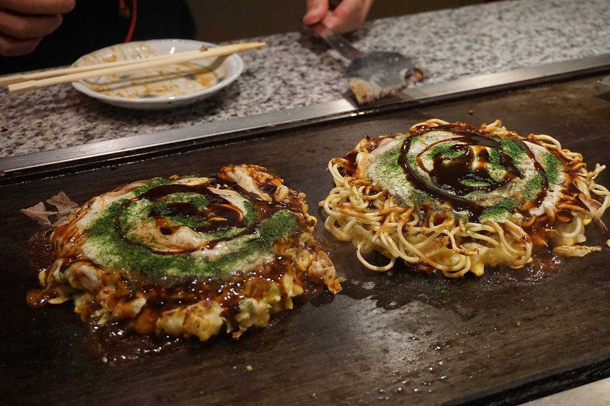To okonomiyaki som steker på en teppanplate, dekket av brun saus i sikksakk og grønt tangpulver
