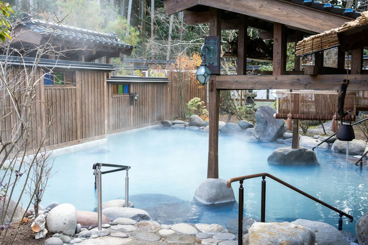 Tomt utendørsbad med melkeblått, dampende vann omgitt av runde steinblokker, trepanel og hengelykt i en japansk hage