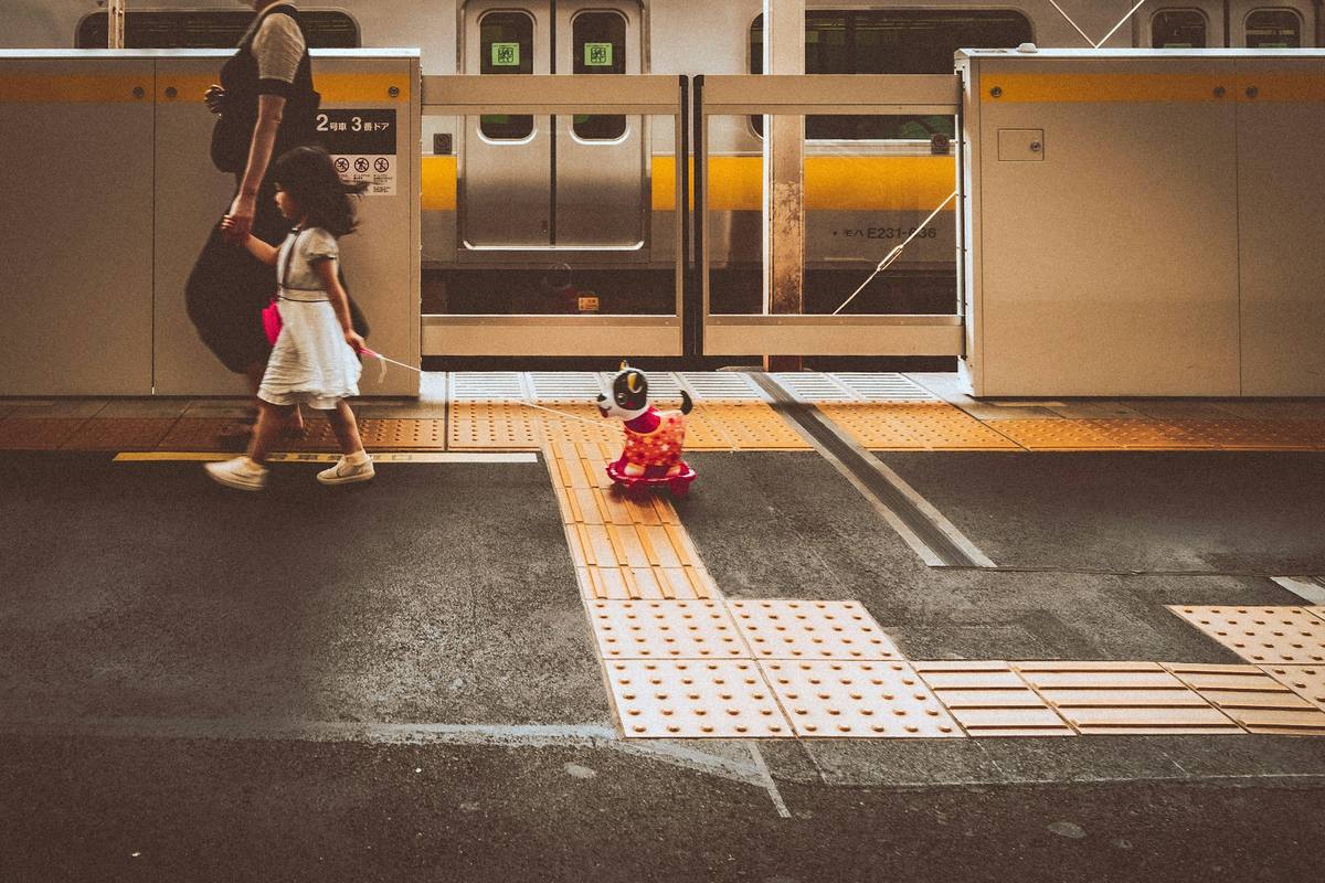 En voksen holder en liten jente i hånden på en japansk perrong mens hun trekker et leketøy på hjul etter seg, foran et lokaltog med gul stripe og perrongdører merket med vognnummer på japansk