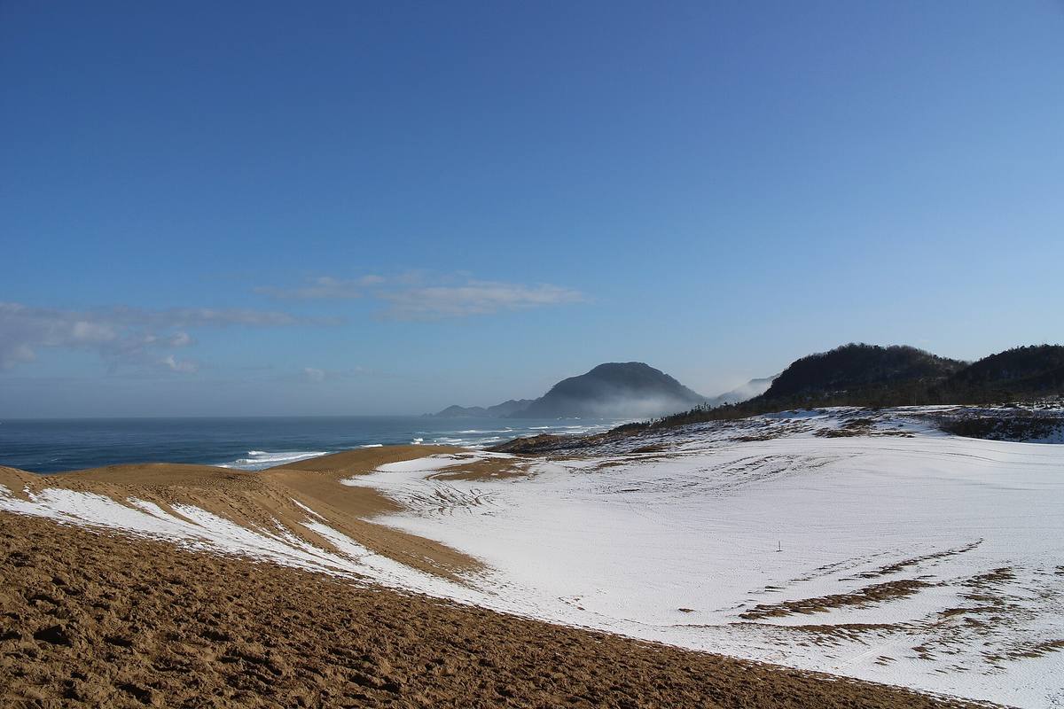 Sanddynene i Tottori delvis dekket av snø, med havet og mørke fjell i bakgrunnen
