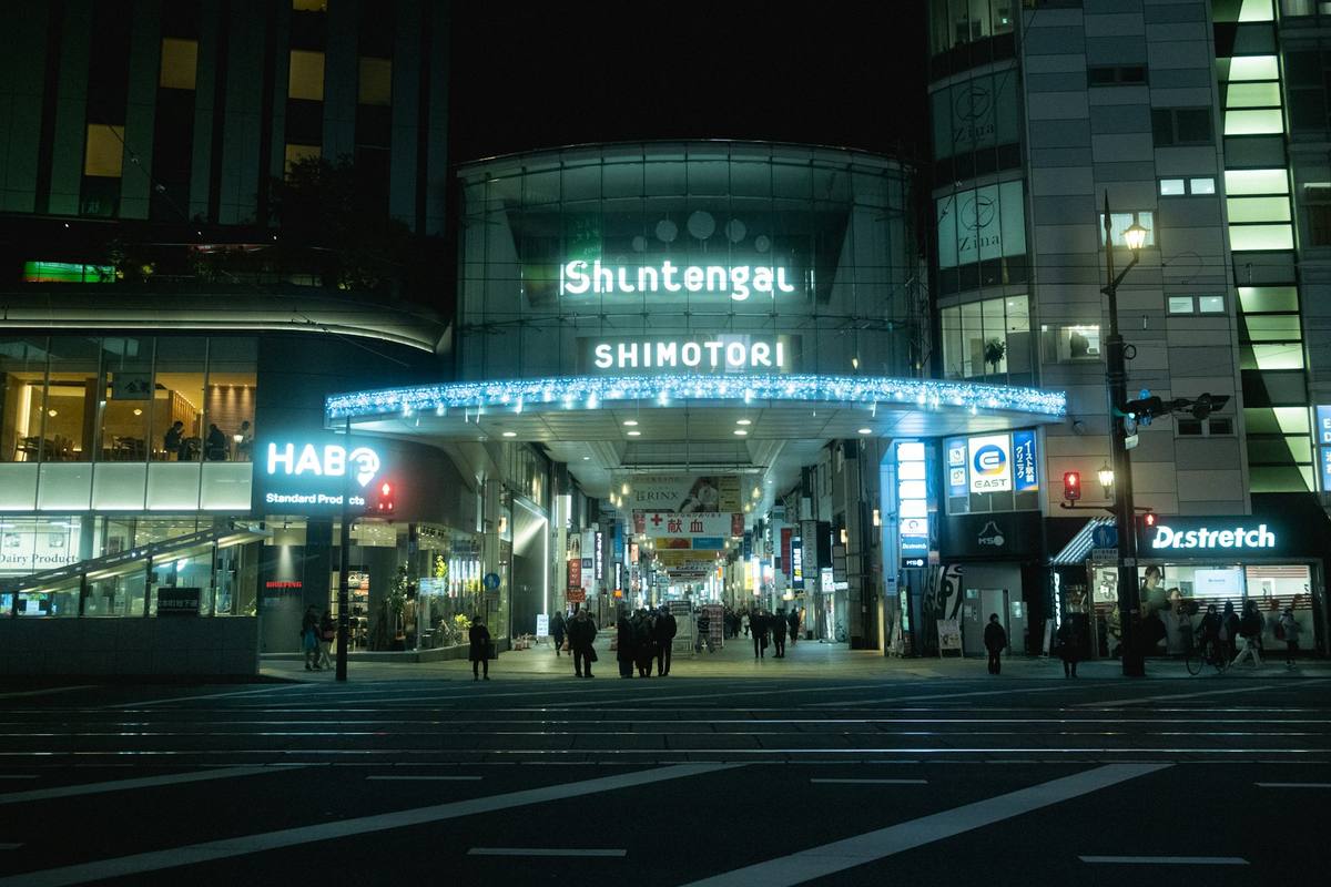 Opplyst inngangsparti til en overbygd handlegate om kvelden, med lysende buegang over inngangen og butikkskilt i flere etasjer på begge sider