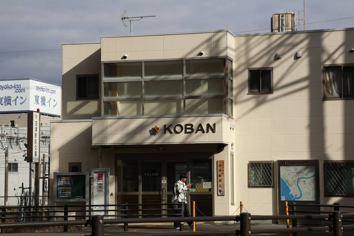 Lys politipost med skiltet KOBAN over inngangen, oppslagstavle ved siden av og et områdekart på veggen til høyre, fotografert i dagslys