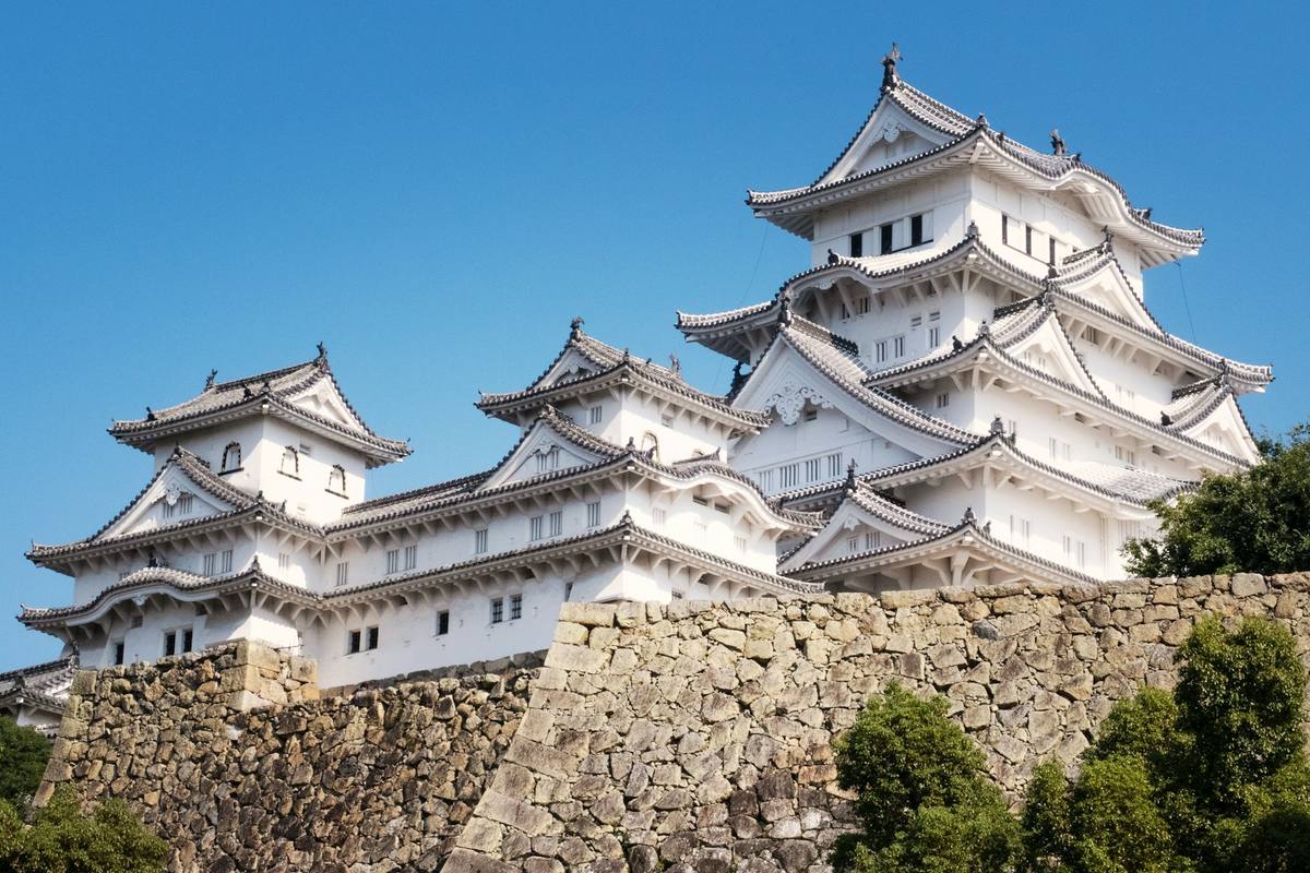 Himeji-slottets hvitkalkede borgtårn med buede takgavler i flere etasjer over en massiv steinmur mot dyp blå himmel