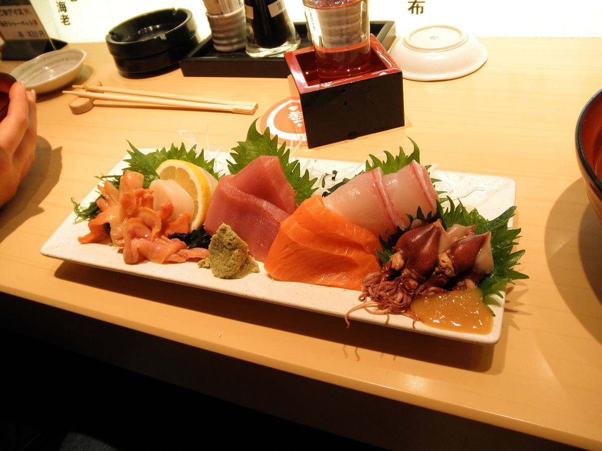 Avlangt fat med rå fiskeskiver i rødt, oransje og hvitt lagt på grønne shiso-blader, med en klatt wasabi og en sitronskive ved siden av