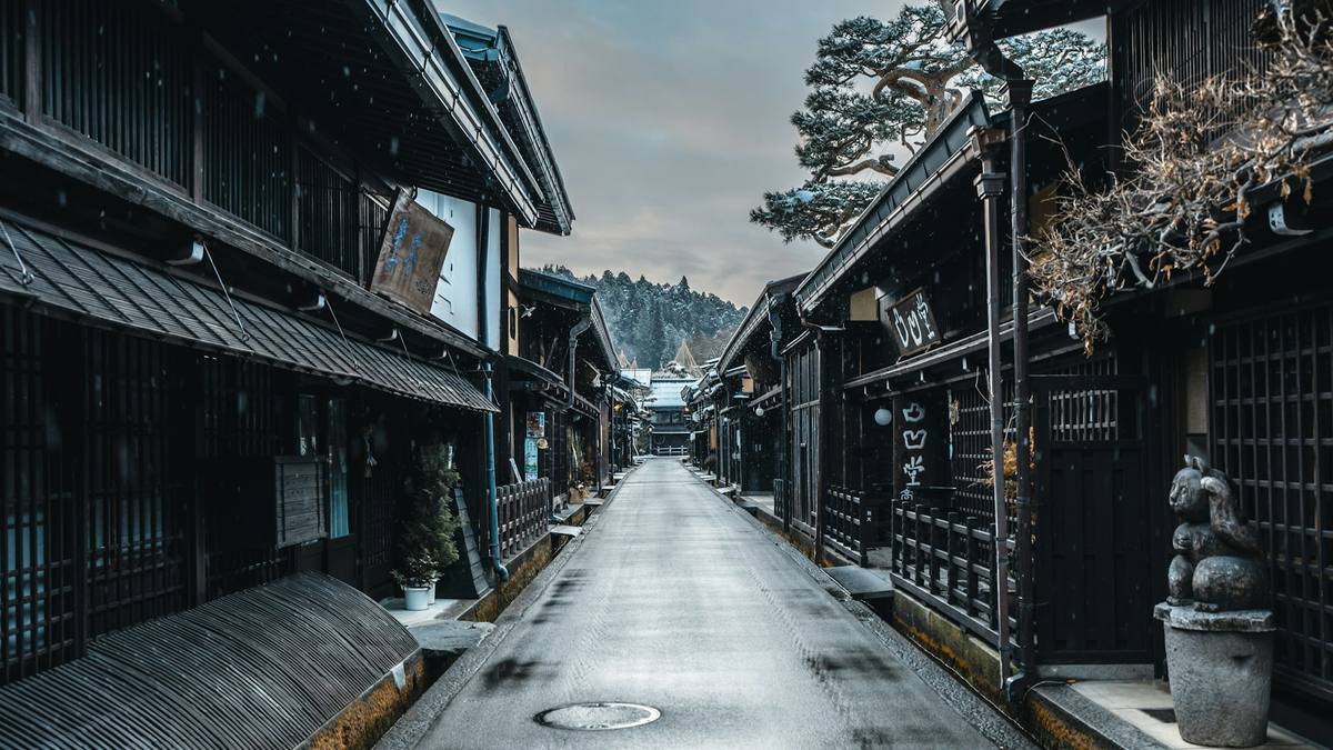 Smal gate mellom to rekker mørke trehus med gitterverk foran vinduene, lett snøfall og skogkledde fjell i enden av gata
