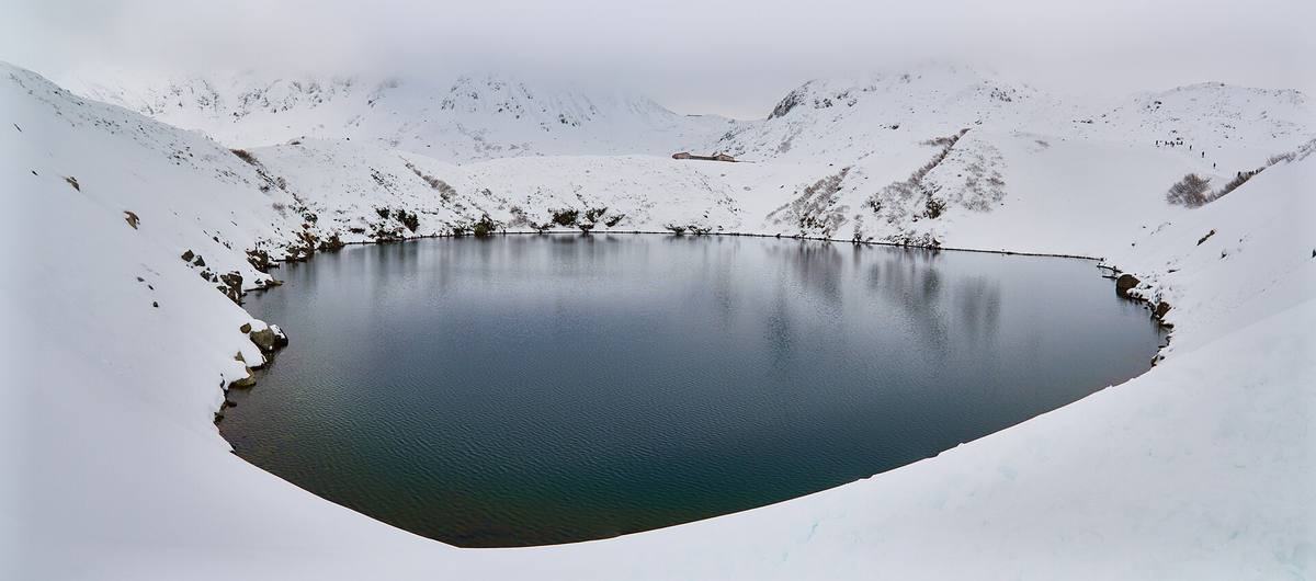 Det mørke kratersjøvannet i Mikurigaike ligger stille midt i et snødekt høyfjellsplatå med hvite topper i bakgrunnen