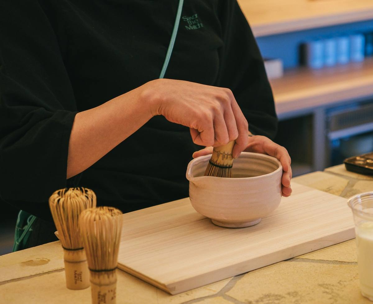 Hender som visper matcha med en bambusvisp i en lys keramikkskål, med flere visper stilt opp på benken ved siden av
