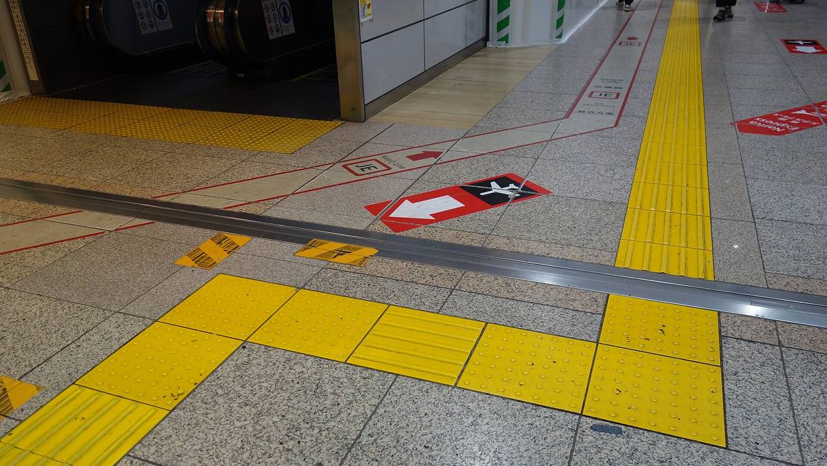 Gule ledelinjer med knotter og riller støpt inn i gulvet på en japansk togstasjon, sammen med røde piler og markeringer for påstigning
