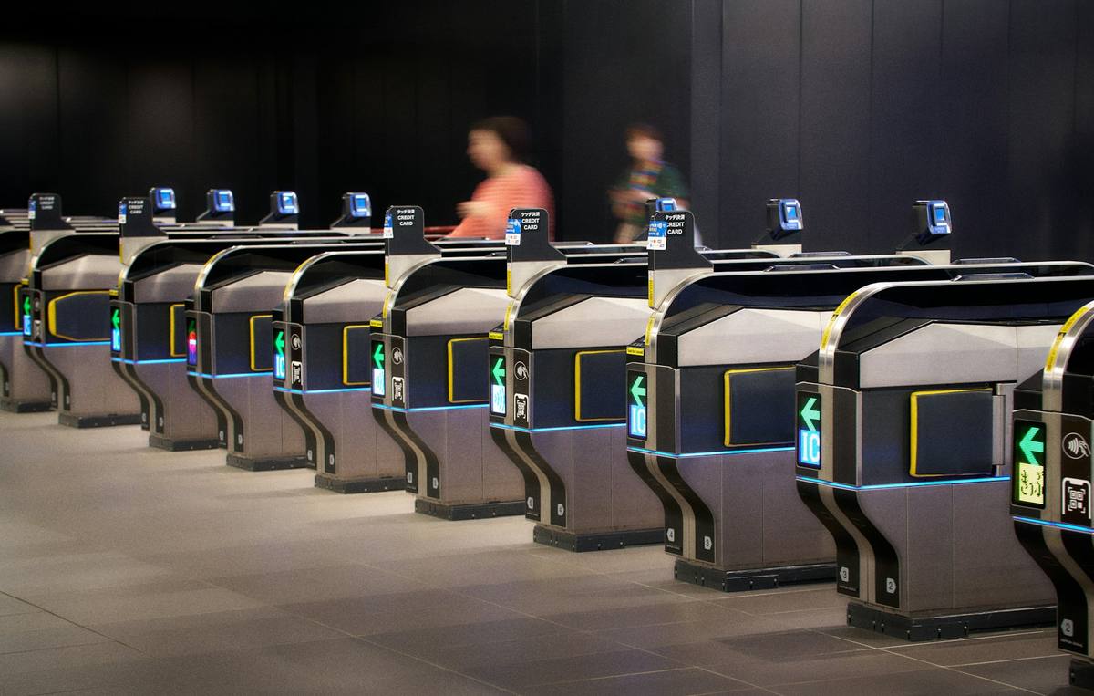 Rekke av automatiske billettsluser i en stasjon med grønne piler, IC-lesere og merker for kontaktløs betaling, mens to reisende passerer i bakgrunnen