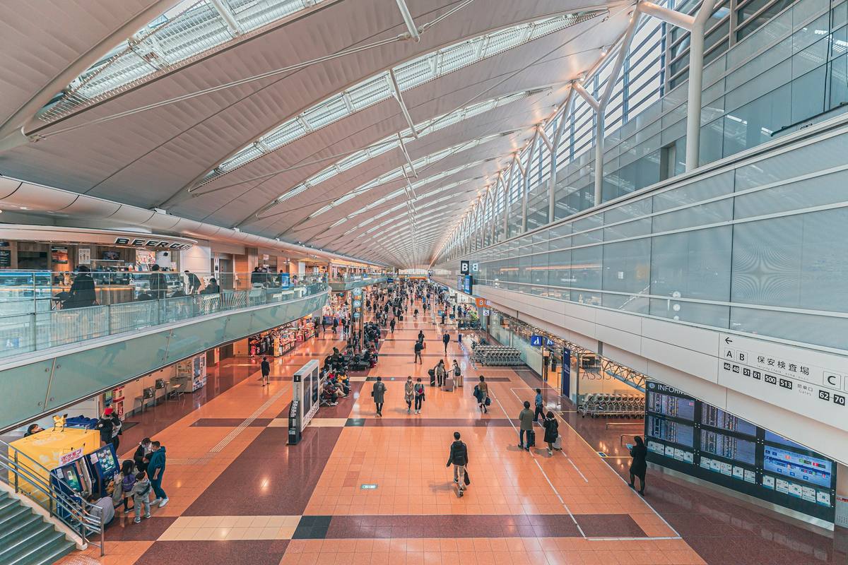 Lang terminalhall på en japansk flyplass sett ovenfra, med buet glasstak, reisende med koffert og skilt til sikkerhetskontroll og gater på japansk og engelsk