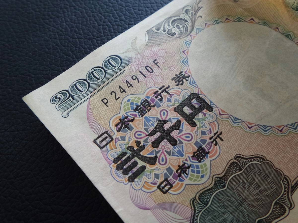Nærbilde av hjørnet på en japansk 2000-yenseddel som ligger skrått på en mørk skinnflate, med tallet 2000 og Japans sentralbanks navn i sort skrift over et mangefarget mønster