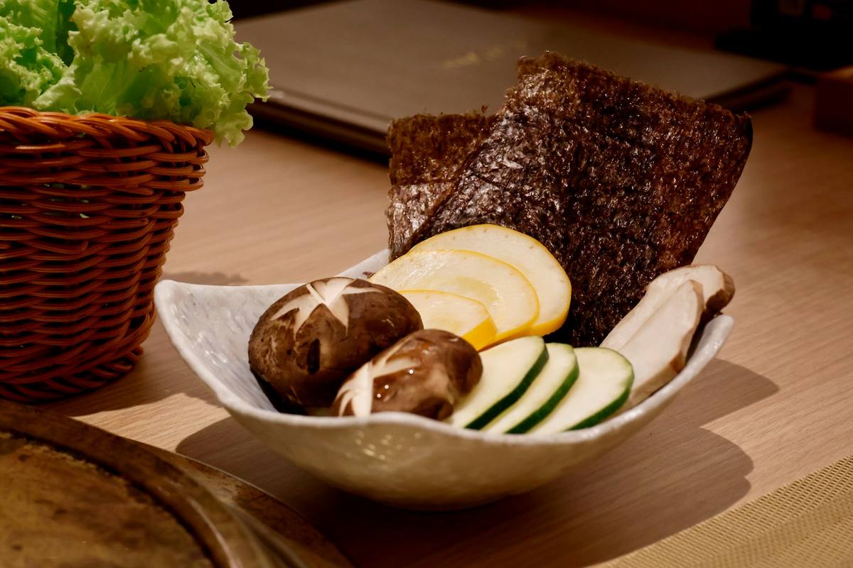Keramikkskål med shiitakesopp, skiver av squash og agurk og et ark tørket nori, med en kurv med salat bak