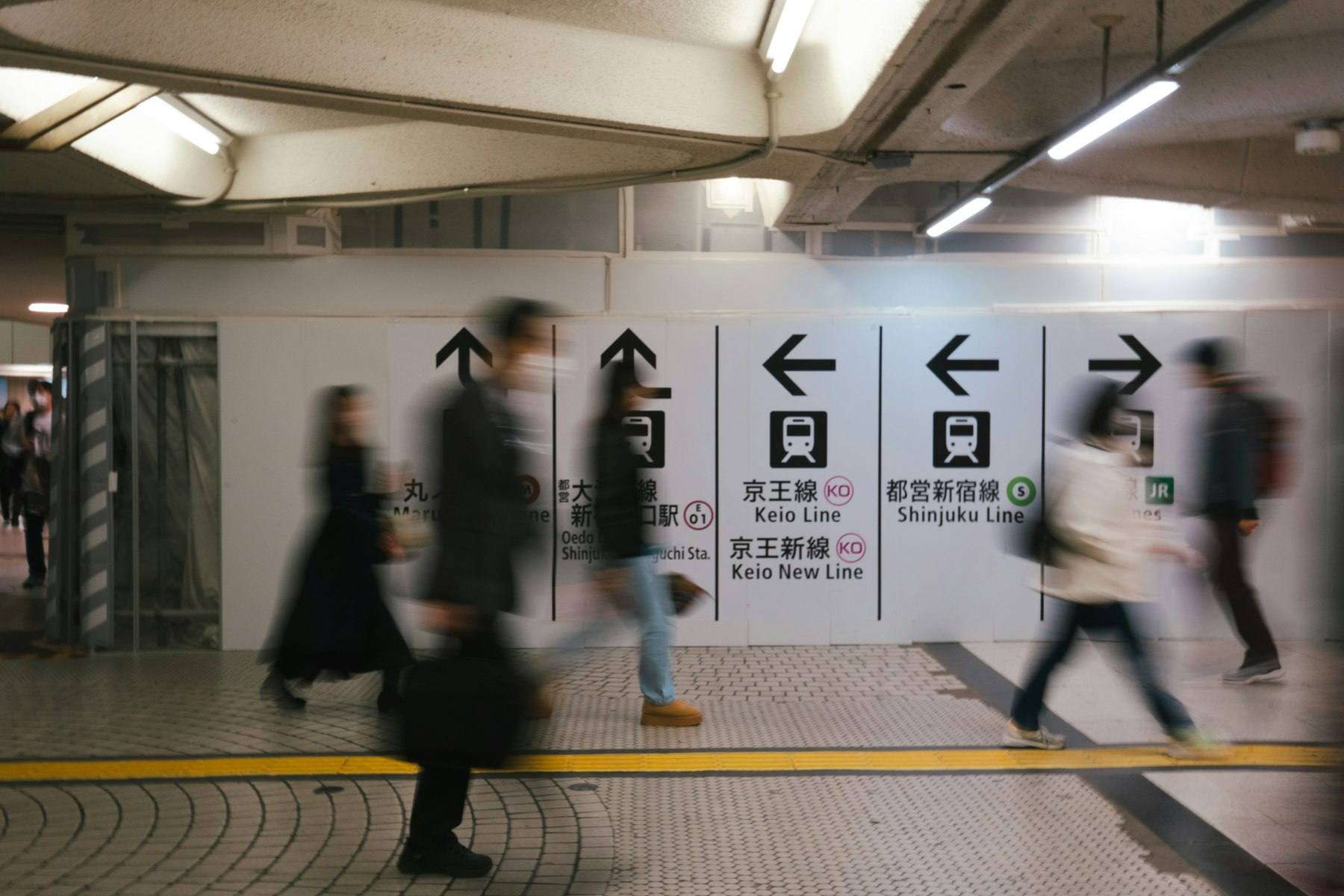 Skiltvegg i en undergrunnsstasjon med piler og linjenavn på japansk og engelsk mens pendlere passerer i bevegelsesuskarphet